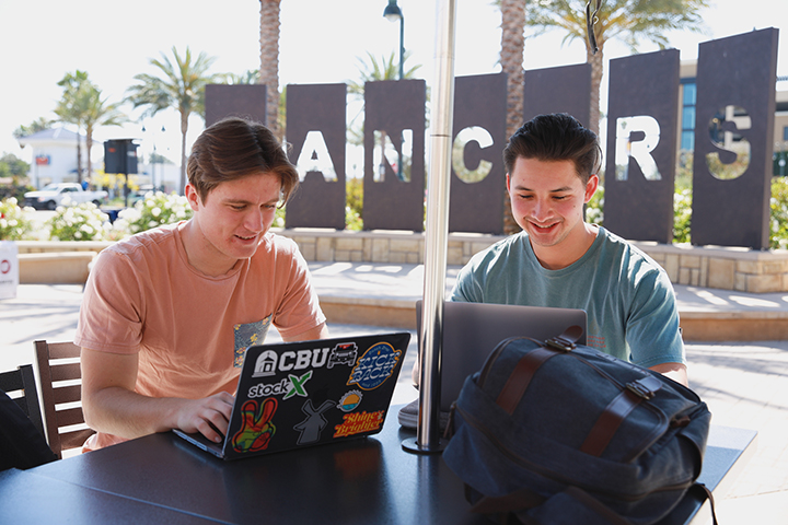Two students sit at an outdoor table while working on laptops