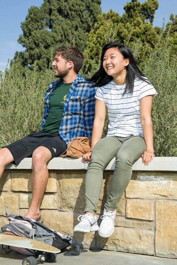 Two CBU students sitting on a stone wall by a sidewalk on campus
