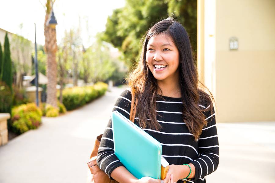 A smiling female international CBU student holding books and folders while walking down a sidewalk on campus